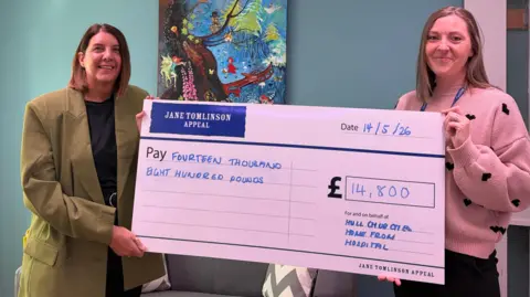 Two people are standing indoors against a blue-green wall, holding a large ceremonial cheque from the “Jane Tomlinson Appeal.” The cheque is dated “14/5/26” and made out for “Fourteen thousand eight hundred pounds,” with the amount “£14,800” written in a large box on the right. Beneath the amount, handwritten text reads “For and on behalf of Hull and Ousea Home From Hospital” 
