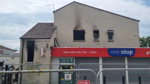 The burnt out windows of first floor flat can be seen in the images and a one stop shop with its shutters down.