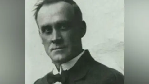 A black and white image of David Danskin. A clean-shaven man with short black hair is wearing a black bow-tie, white shirt and a black blazer 