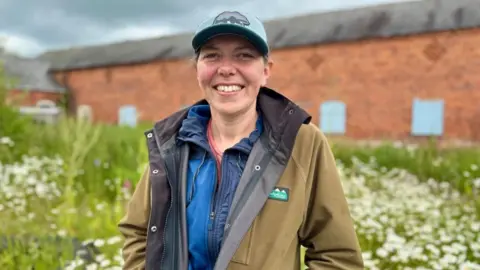 A woman standing in front of a red brick farm building. She is wearing a khaki coat and a blue cap. She is smiling and there is a short, grass meadow behind her. 