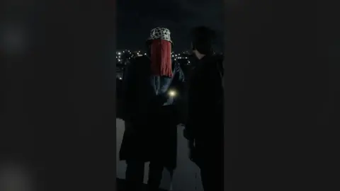 A man wearing a Dalmatian print hat with red tassels hanging from the edges to cover his face stands on a rooftop next to another man. Both are dressed in black.