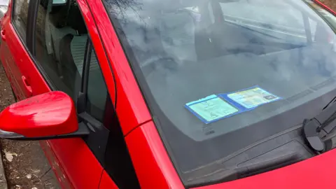 A red saloon car in a car park, with a blue badge displayed prominently on the dashboard.