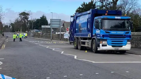 Blue garbage truck in police cordon on empty street. Two officers wearing safety vests can be seen in the distance.