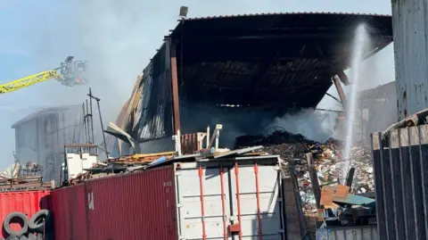 Michael Nagasaka/BBC An industrial area with a large pile of rubbish with a metal roof and other metal units next to it. Smoke can be seen rising around it along with water being sprayed high up into the air.