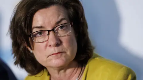First Minister Eluned Morgan is looking past the camera and her head turned, wearing a yellow jacket and glasses.