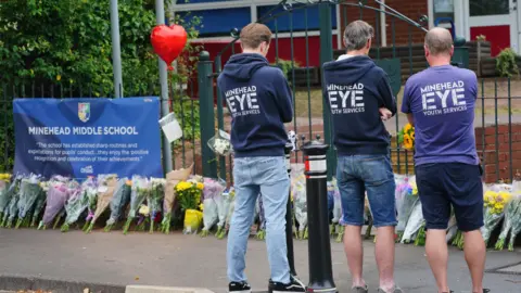 Three men stood with jumper's reading 'Minehead Eye' have their backs to the camera, looking at a row of flowers which have been laid in front of a fence. A sign saying 'Minehead Middle School' is attached to the railings.