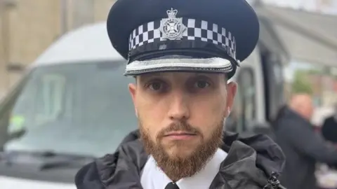 A policeman with a neatly clipped light brown beard stares at the camera with a neutral expression. He is wearing a black police jacket over a white shirt and a police hat.