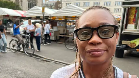Mousumi Bakshi/BBC A woman - Milli Akehurst - looking at the camera with a neutral expression. She is wearing sunglasses and standing in front of a market, with a group of people behind her.
