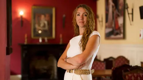 Samantha Harper, a woman with long curly blond hair, stands with her arms folded in the lounge of a historic pub with oil portraits hanging on the wall. 