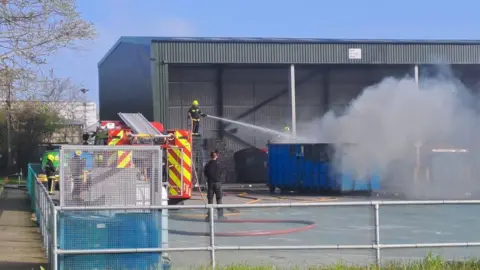 Somerset Council A firefighter in a yellow helmet stands on a tall platform holding a powerful water hose, which is being directed into a large blue metal skip. The skip is emitting large clouds of white smoke. Next to the firefighter is a red and yellow striped fire engine. Behind is a large open metal-panelled building.