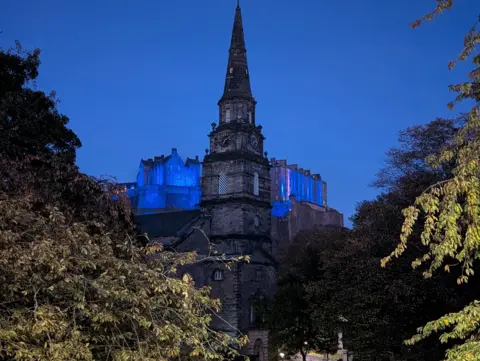 Alex Orr St Cuthbert church in Edinburgh in the foreground with Edinburgh Castle in the background, illuminated in blue lights below a clear, blue sky in the evening