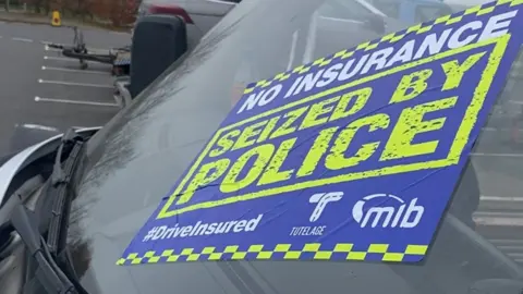 A close-up of a blue sticker on a car windscreen. It says no insurance in white letters and seized by police in yellow letters