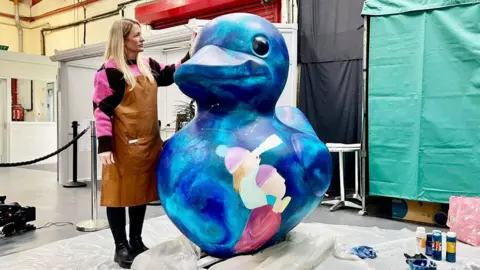 BBC A woman in a brown apron is stood beside a 1.5m high duck statue that has been decoratively painted in a marketplace entrance. The woman has long blonde hair, a pink and black jumper, black leggings and boots. The duck is painted in blue and green swirls with white constellations and stars also painted on. There is a painted image of a man with a child on his shoulders looking through a telescope. The duck is stood on a pallet and white ground sheets. There are bottles of paint beside it. 