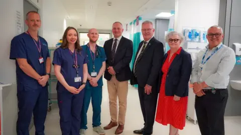 Department of Health Mike Nesbitt has grey hair and glasses and is wearing a dark suit while standing among hospital staff, some who are in blue scrubs. They are standing on a hospital ward.