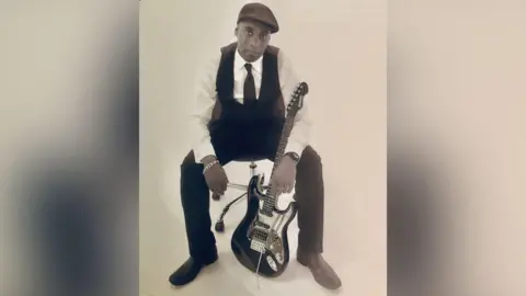 Pauline Reid A sepia toned photo of Paul Reid sitting on a chair. He is wearing a flat cap, white shirt, black tie and black waistcoat with black trousers and shoes. He is looking into the camera and has a black and white guitar leaning on him.