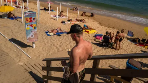 Getty Images A man with a black cap and no shirt on walks down to a beach in Albufeira, where other people are lying on the yellow sand.