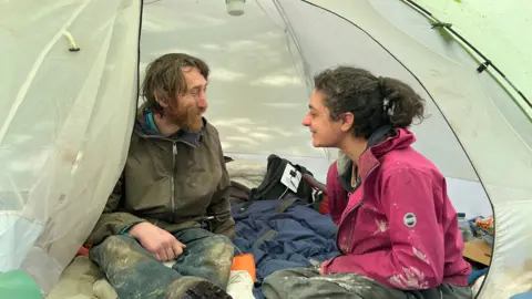 A man and woman sit inside a white tent. The man has long brown hair, a brown beard, a green coat and blue jeans. The woman has black hair tied back, a navy hoodie and a pink coat. There are sleeping bags and clothes around them.