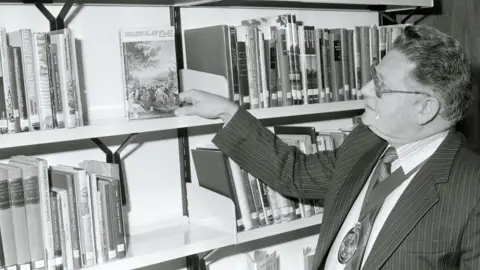 BANES Council A black and white picture of a man wearing a pinstripe suit and glasses. He is looking at a book on a shelf admiringly, while other books are stacked beside it on the shelves using a dewey decimal system. 