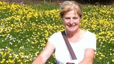 Danielle smiles at the camera in a grainy photograph. She has blonde hair and wears a white t-shirt. She is sitting in a meadow or garden with long grass and lots of daisies and buttercups.