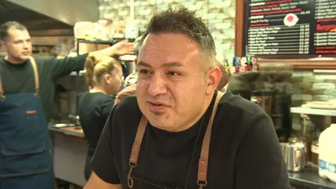 A man speaks off from the camera, wearing a black t-shirt and brown overalls standing behind a café counter 