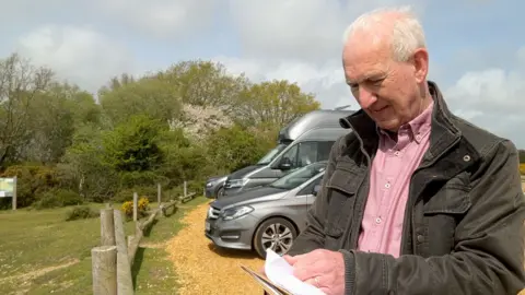 Nick Hubbard is wearing a brown jacket and pink shirt and has short grey hair. He is looking down at his notes on a clipboard while standing in a car park. Behind him are several parked cars, a wooden fence and trees and bushes. 