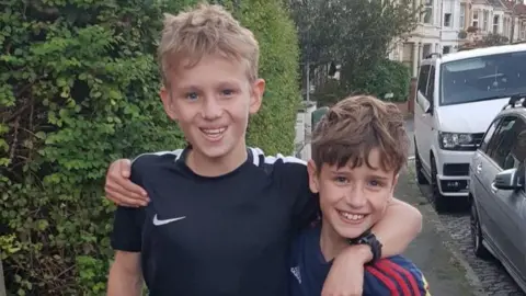 Handout George (left) and Artie (right) smile at the camera with their arms around each other's shoulders. George has blonde hair and wears a black Nike t-shirt, while Artie has brown hair and wears a red and blue striped football shirt. 