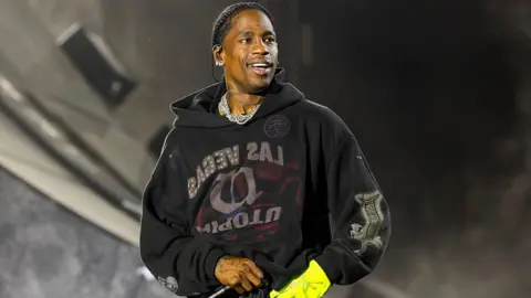 Travis Scott performs on stage wearing a black sweatshirt and a yellow glove. 