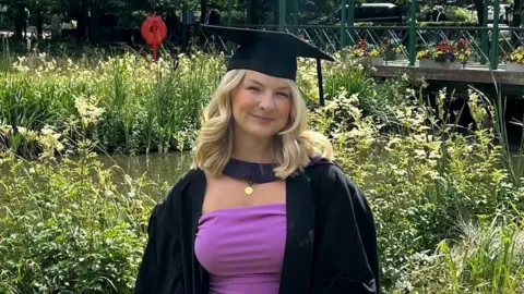 Charlotte Gill A blonde woman wearing a graduation cap and gown smiles as she stands by a pond in a park