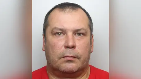 Dariusz Meczynski mug shot. He has short brown hair and is wearing a red t-shirt. The background is neutral grey.