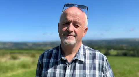 John Davies, a man in roughly his 60s, with a short white goatee and short white hair. He has sunglasses on his head and is wearing a chequered shirt with lots of different shades of blue