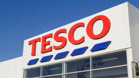 A Tesco logo on a white building. There is a blue sky above. 