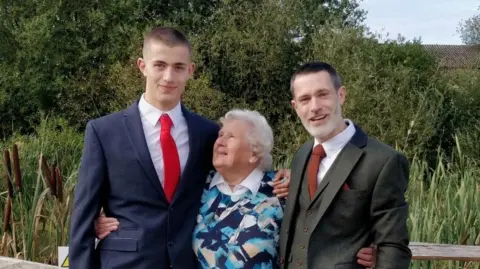 Jane Radford Will Radford is on the left with short dark hair wearing a navy blue suit with a white shirt and red tie. In the middle, his grandmother laughs as she looks up at him. She has white curly hair and wears a patterned jumper over a white shirt. On the right Will's father has greying hair and wears a grey suit and waistcoat with a white shirt and brown tie.