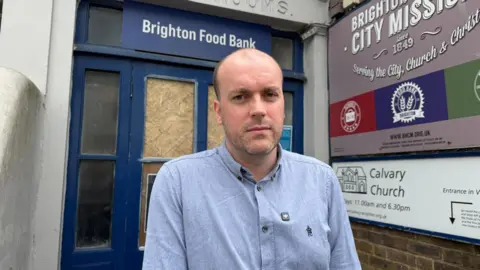 A bald man in a pale blue shirt, standing in front of a navy blue and glass door which has the words Brighton Food Bank on it in white