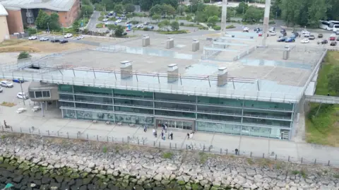 BBC/Jon Coates An aerial view of the National Glass Centre which shows a large glass fronted building with a glass-topped roof, which visitors used to be able to walk on. The building also has seven large turrets which help support the overall structure, and in the background are two large chimneys.