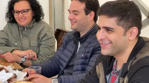 BBC A woman and two men sit at a table inside a tent smiling as they have some food and drink