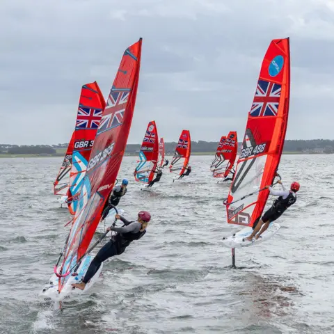 Mike Pringuer A series of wind surfers with bright red sails carrying the Union Jack