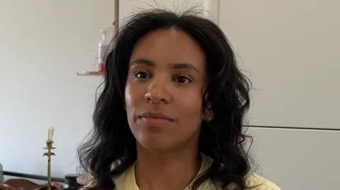 A woman with long curly black hair, wearing a yellow T-shirt, sits in front of a cupboard. She is looking away from the camera.