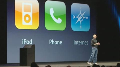 Steve Jobs in his black polo neck, blue jeans and white trainers, standing onstage in front of a display screen, with enlarged logos for iPod, Phone and Internet behind him.  The iPod log s a graphic of the device on top of an orange background, the Phone icon has a picture of a phone on a green background and 'Internet' has a compass on top of a blue background.  A podium is visible to the right hand side of the stage.