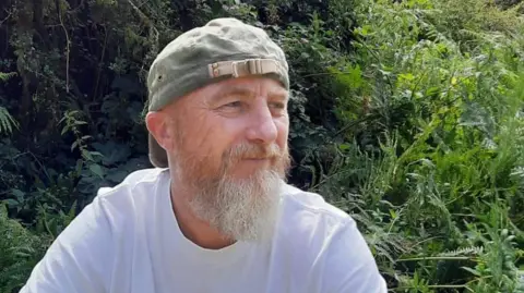 Mark Page Mark Page. He has a white/grey beard and is looking to the right of the picture. He is wearing a white shirt and khaki-coloured baseball cap. Behind him are green shrubs and foliage.