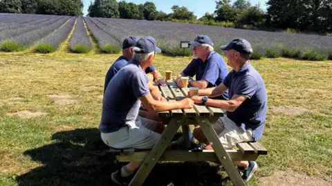 A group of men sit at a wooden picnic bench on grass, with a field of lavender behind. They are all wearing navy t-shirts and baseball caps and have paper cups front of them as they talk.
