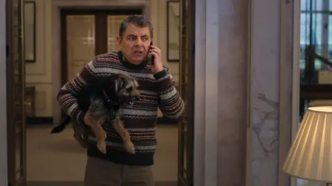 Netflix Rowan Atkinson stands inside a very posh house with marble walls and hotel style lamps. He holds the dog under one arm and is talking on a mobile phone, with an irritated expression on his face. He wears a Fairisle style knitted jumper. Artie has a collar on and his tongue is partly hanging out.