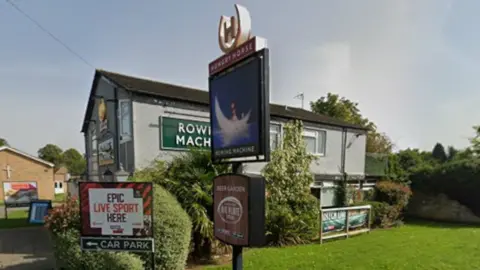 The Rowing Machine pub is a relatively compact building in front of a lawn on a street corner, with signs outside advertising the pub and TV sports coverage.