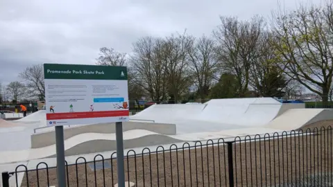 A concrete skate park. The concrete is white and cream. There are trees to the right. It is cloudy above. In the foreground is a dark-painted fence and a sign reading PROMENADE PARK SKATE PARK.