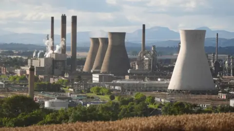 PA Media A general view, taken from a high hilltop, of the Grangemouth industrial complex. Cylindrical concrete structures and chimneys rise out of a network of  pipes and industrial buildings. 