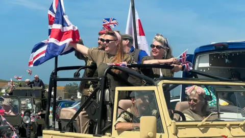 A group of woman with 1940s style hairdos  and wearing khaki coloured clothing and dark glasses stand in the back of a jeep waving the Union Jack on Liberation  Day in May 2024.