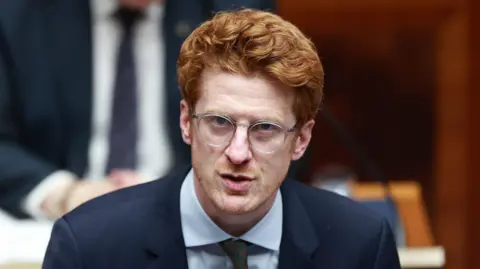 PA Media O'Toole has ginger curly short hair and glasses. He is speaking in the image and is wearing a navy suit jacket, light shirt and dark tie.