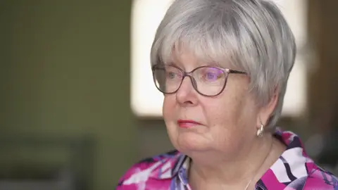 Marion Fellows, who has short grey hair and wears a colourful pink top, looks off camera as she speaks to the BBC.