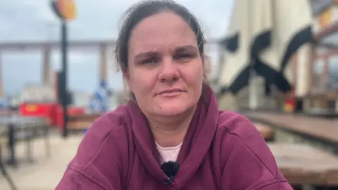 Melanie Watson se sienta en una mesa al aire libre en un café. Lleva una tapa con capucha morada sobre una camiseta de color rosa pálido y tiene su cabello castaño atado hacia atrás. 