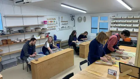 Contributed Six people stand or sit behind pottery stations. Four people sit behind pottery kilns working on clay. Two others stand behind a table and roll out clay for their creations. Pottery equipment rests on walls, shelves and in drawers. 