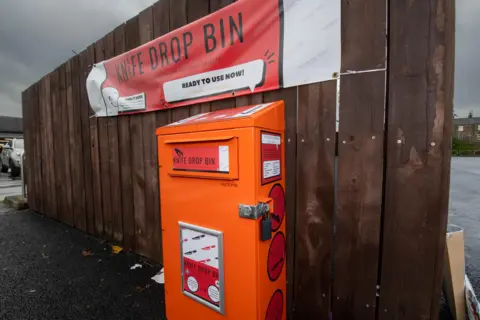 Knife bin with a padlock on it, in front of a wooden fence